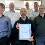 A group of people stands together and smiles for the camera. A man at center holds a framed certificate from BSI in 2023 for ISO 50001.